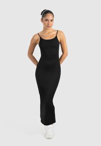 Schwarzes, körperbetontes Spaghetti-Trägerkleid mit einer glatten, gerippten Textur, das bis zu den Knöcheln reicht, kombiniert mit weißen Sportschuhen.