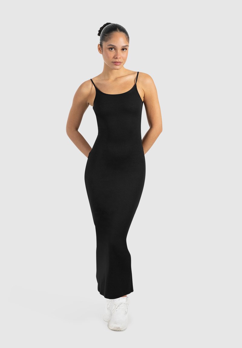 Schwarzes, körperbetontes Spaghetti-Trägerkleid mit einer glatten, gerippten Textur, das bis zu den Knöcheln reicht, kombiniert mit weißen Sportschuhen.