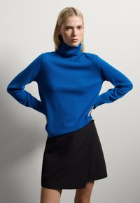 Maglione a collo alto blu in materiale morbido, dotato di maniche lunghe e una vestibilità morbida, abbinato a una gonna avvolgente nera.