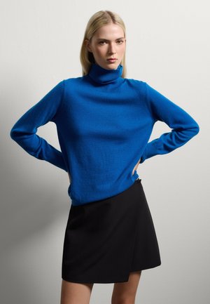 Blauwe coltrui van zacht materiaal, met lange mouwen en een losse pasvorm, gecombineerd met een zwarte wikkelrok.
