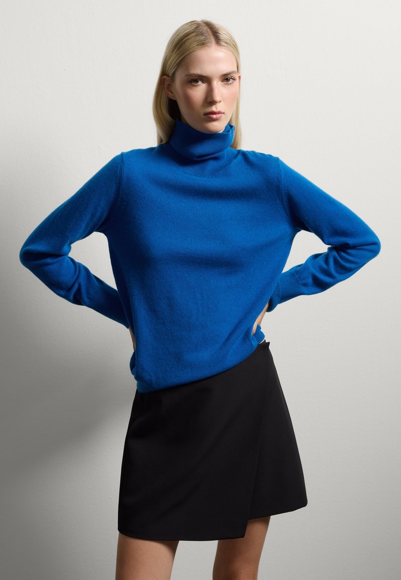 Maglione a collo alto blu in materiale morbido, dotato di maniche lunghe e una vestibilità morbida, abbinato a una gonna avvolgente nera.