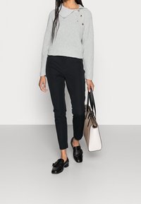 Maglione grigio chiaro a maglia con colletto ampio e bottoni, abbinato a pantaloni neri slim-fit e mocassini neri. Tiene in mano una borsa strutturata beige.
