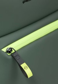 Borsa in tessuto impermeabile verde scuro con una zip gialla brillante. Presenta un cursore della zip nero e un'etichetta con un logo. Texture liscia.