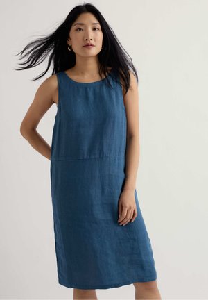 Femme aux longs cheveux noirs portant une robe bleue sans manches jusqu'aux genoux, debout devant un fond clair uni.