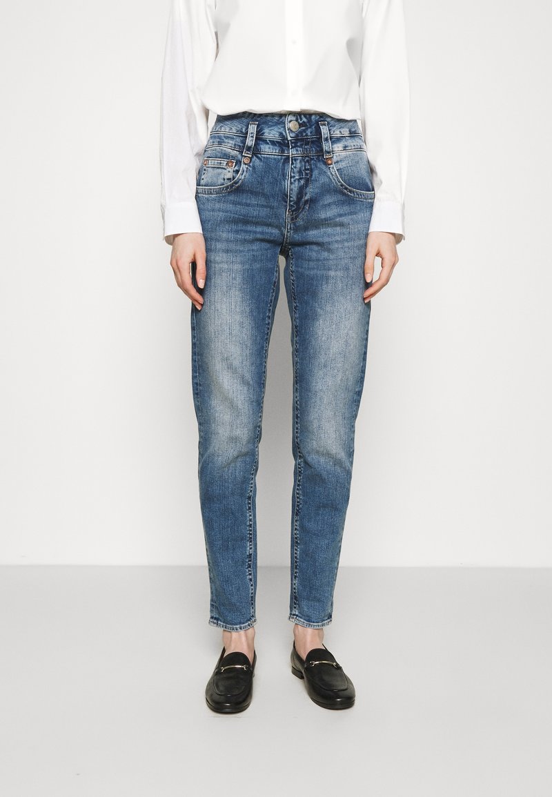Jean en denim bleu clair avec une coupe droite, finition délavée, cinq poches et fermeture à bouton. Porté avec une chemise blanche à manches longues et des mocassins noirs.