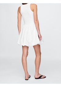 Robe blanche sans manches avec un haut ajusté et une jupe évasée et froncée. La texture du tissu semble lisse, avec un design superposé et volumineux.