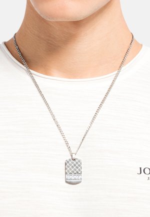 Collarín de perro de plata con una superficie superior texturizada, exhibido en una cadena, usado sobre una camiseta de color claro.