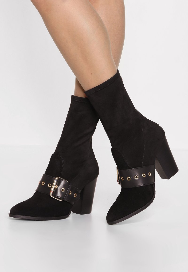 Bottines en daim noir avec talons épais et larges sangles en cuir décoratives comportant des œillets et boucles dorés, portées sur des jambes croisées.