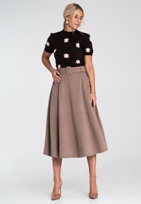 Kobieta w czarnej bluzce z krótkim rękawem w różowe kwiaty oraz w beżowej midi spódnicy z wysokim stanem i paskiem, nosząca cieliste szpilki i kolczyki.