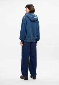 Μπλε υπερμεγέθη φούτερ με κουκούλα και κορδόνι, ελαφρύτερη εσωτερική επένδυση, ριμπ μακριά μανίκια και κεντημένο σχέδιο. Συνδυασμένο με φαρδιά τζιν παντελόνια.