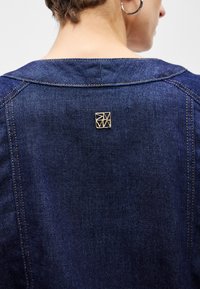 Τζάκετ τζιν με σκούρο χρώμα, με χρυσά γεωμετρικά αξεσουάρ στην πλάτη του λαιμού και αντίθετες ραφές.