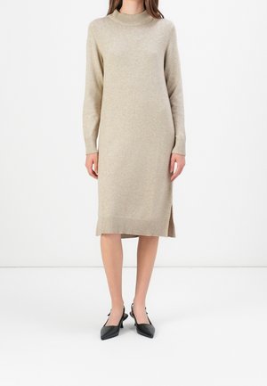 Femme portant une robe midi en maille beige à manches longues avec un col montant et une fente latérale, associée à des escarpins noirs à bouts pointus et bride arrière.