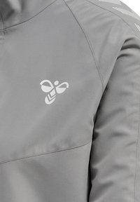 Szara kurtka z białym logo w kształcie pszczoły na piersi oraz widocznymi szwami na ramieniu i rękawie.