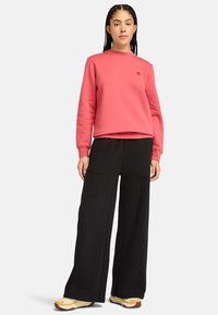 Rosa sweatshirt med rund halsringning och broderad logotyp, kombinerad med svarta vida byxor och beige skor med texturerade sulor.