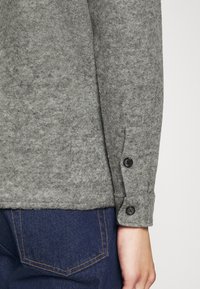 Chemise en feutre gris à manches longues, avec deux boutons noirs au poignet. Le tissu a une texture douce et une coupe nette et ajustée.