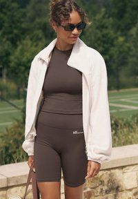 Barna testhezálló aktív viselet felső és rövidnadrág, fehér, cipzáras, polár dzsekivel párosítva. A modell sötét napszemüveget visel és táskát tart.