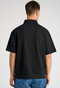 Czarna koszulka polo z krótkim rękawem o luźnym kroju, z stójką i przeszytymi detalami na krawędziach, w zestawie z niebieskimi dżinsami.