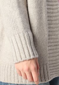 Lys grå strikket cardigan med ribbestrikkede mansjetter og kant. Myk tekstur, middels tykkelse, som viser en avslappet passform og koselig utseende.