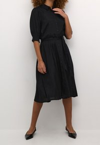 Robe noire à boutons avec des manches courtes bouffantes, taille ceinturée et jupe à hauteur des genoux. Tissu lisse avec un léger éclat. Portée avec des chaussures plates noires.