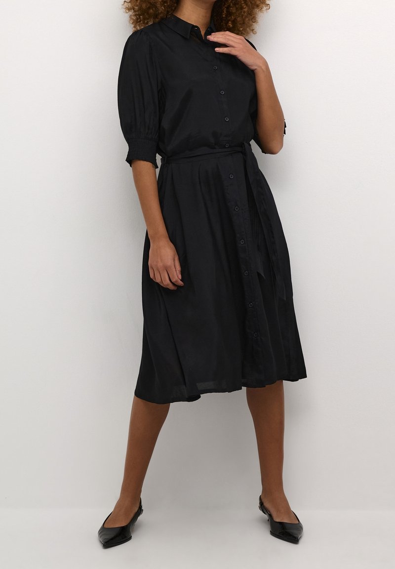 Robe noire à boutons avec des manches courtes bouffantes, taille ceinturée et jupe à hauteur des genoux. Tissu lisse avec un léger éclat. Portée avec des chaussures plates noires.