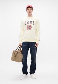 Kremowy sweatshirt z niebieskim i czerwonym logo, ciemnoniebieskie dżinsy, białe sneakersy oraz beżowa torba z brązowymi akcentami. Modelka nosi różową czapkę.