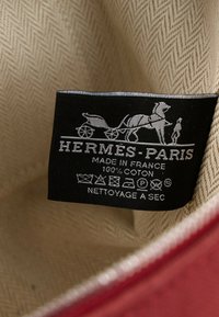 Étiquette en tissu Hermès-Paris noire avec texte blanc et logo de calèche tirée par des chevaux sur tissu beige texturé et bordure de fermeture éclair rouge.