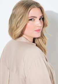 Blusa beige leggera con una consistenza morbida, caratterizzata da un colletto semplice e un dettaglio di piega sul retro. La modella ha i capelli lunghi e mossi.