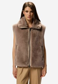 Prsni brezrokavnik iz umetnega krzna v mehki rjavi barvi, z visokim ovratnikom, sprednjim zadrgo in brez rokavov. Gladke teksture z bočnimi žepi.