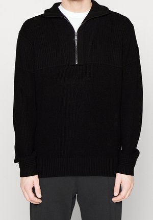 Pull-over en tricot noir avec un col zippé demi-long, présentant une texture côtelée. Manches longues et poignets ajustés, mettant en avant une silhouette décontractée.