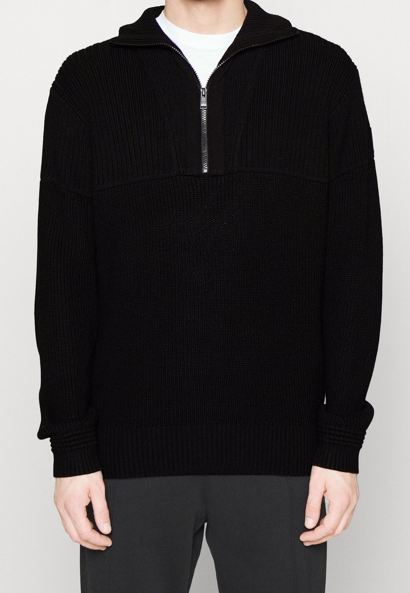Pull-over en tricot noir avec un col zippé demi-long, présentant une texture côtelée. Manches longues et poignets ajustés, mettant en avant une silhouette décontractée.