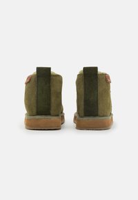 Para oliwkowych zamszowych klapek z brązowymi podeszwami i ciemnozielonymi pionowymi językami, pokazana z tyłu na białym tle.