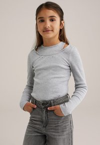 Hellgraues, langärmeliges, geripptes Shirt mit Rundhalsauschnitt, verziert mit kleinen dekorativen Nieten und einem taillierten Schnitt, kombiniert mit hochgeschnittenen grauen Jeans.