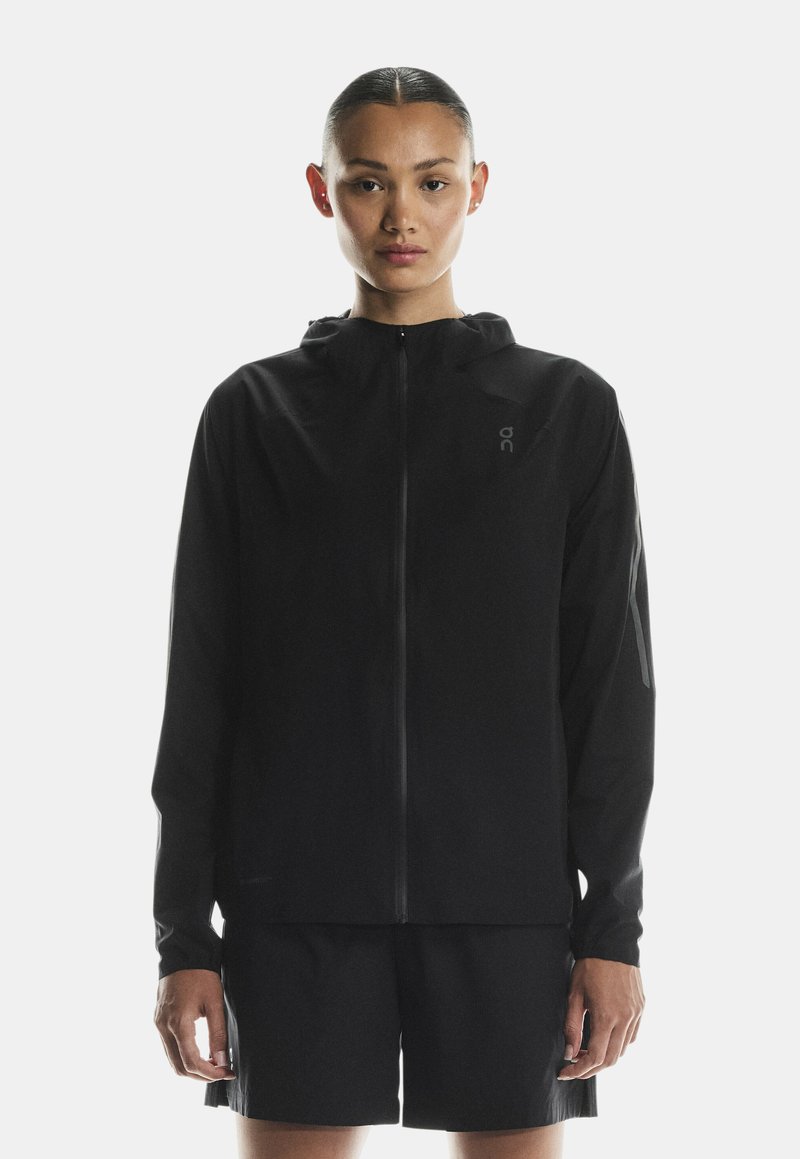 Juoda, lengva striukė su pilnu užtrauktuku, gobtuvu ir minimalistikos dizainu. Yra subtilus prekės ženklas ir glotni, lygi tekstūra.