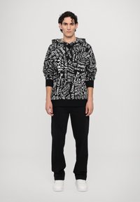 Hoodie présentant un motif de texte abstrait en noir et blanc, avec une capuche à cordon, des poignets côtelés et une coupe décontractée. Associé à un pantalon noir.