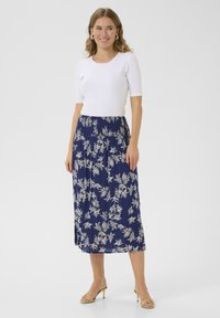 Jupe midi florale navy avec des motifs de feuilles blanches et une taille smockée, associée à un haut blanc ajusté et des sandales à talons dorés.