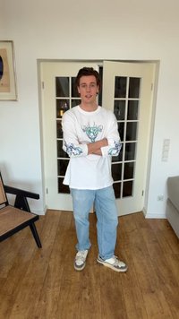 Νέος άνδρας με σταυρωμένα χέρια, φορώντας λευκό πουκάμισο με μακριά μανίκια και πράσινα σχέδια, ανοιχτόγαλαντζια τζιν και αθλητικά παπούτσια, στέκεται σε εσωτερικό χώρο πάνω σε ξύλινο πάτωμα.