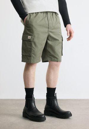 Homme portant un short cargo olive, des chaussettes Calvin Klein noires et des bottines noires en cuir massif sur un sol gris.