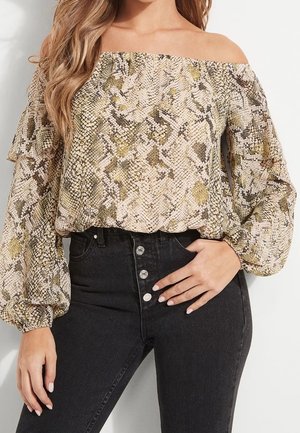 Mujer con blusa de estampado de serpiente sin hombros, mangas largas abullonadas y vaqueros negros de talle alto con botones delanteros.