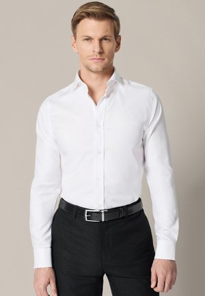 Mężczyzna ubrany w dopasowaną białą koszulę włożoną do czarnych spodni, z czarnym skórzanym paskiem, stojący na jednolitym tle.