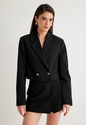 Zwarte cropped blazer met twee knopen, scherpe revers en lange mouwen, gecombineerd met een bijpassende mini-jurk met een diagonale split.