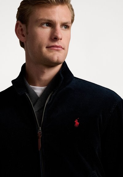 Giacca di velluto a coste blu navy con zip frontale, logo rosa del polo sul petto e colletto nero. Presenta una texture morbida e un design aderente.