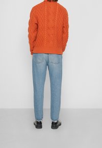 Jersey de punto trenzado en naranja con cuello alto y bajo acanalado, combinado con jeans claros ajustados de color azul y zapatos negros.