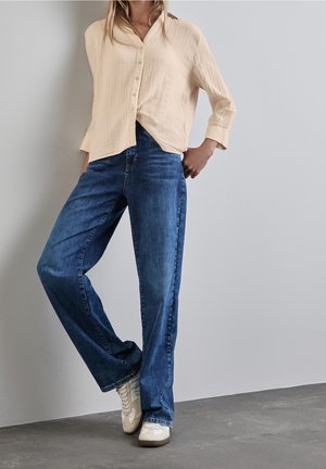 Personne portant une chemise beige à boutons, un jean bleu et des baskets blanches, debout avec une main dans la poche contre un mur uni.