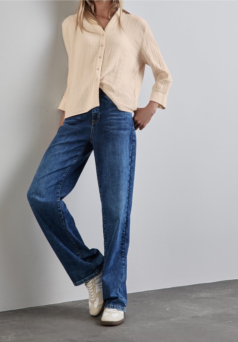 Personne portant une chemise beige à boutons, un jean bleu et des baskets blanches, debout avec une main dans la poche contre un mur uni.