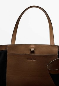 Sac fourre-tout en cuir brun avec des panneaux latéraux noirs, présentant une texture lisse, deux poignées, un fermoir en métal et un logo de marque embossé.