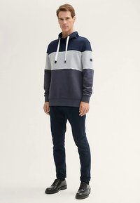 Kapucnis pulóver tengerészkék, szürke és sötétkék színblokkokkal, húzózsinórokkal, elülső kenguru zsebbel és bordázott mandzsettákkal.