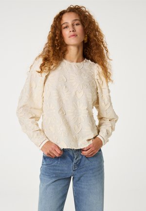 Crème blouse met borduursels in bloemmotieven, met gebloemde lange mouwen en een geschulpte zoom. Gepaarde met lichtblauwe denim jeans.