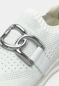 Λευκό πλεκτό sneaker με μεγάλη ασημί μεταλλική πόρπη και ελαστική ζώνη. Διαθέτει περφοραρισμένη υφή και ευέλικτο πάτο.