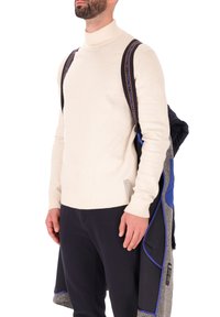 Maglione a collo alto color crema, con polsini e orlo a coste, indossato con una giacca nera con zip caratterizzata da un tessuto grigio e accenti blu, con spallacci dello zaino neri.