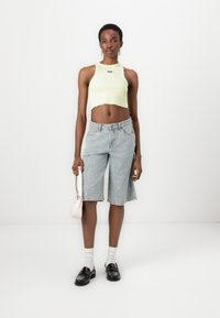 Ljust grönt ribbat crop top med en logotyp, parat med blekta denim Bermuda-shorts och svarta loafers. Håller en liten rosa handväska.